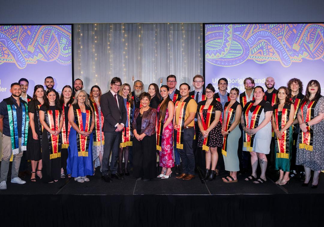 2023 Aboriginal and Torres Strait Islander Sashing Ceremony students pose on stage with Professor Kris Ryan, Deputy Vice-Chancellor (Academic), and Professor Bronwyn Fredericks, Deputy Vice-Chancellor (Indigenous Engagement).